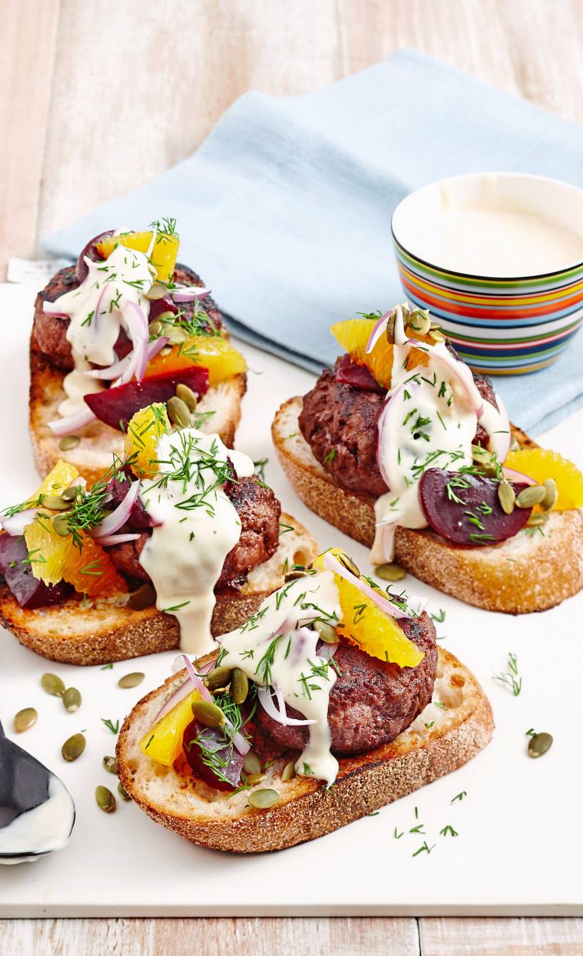 Mehrere geröstete Ciabattascheiben belegt mit Polpette, Roter Bete, Orangenfilets getoppt mit Joghurtsoße und Dill auf einer Servierplatte
