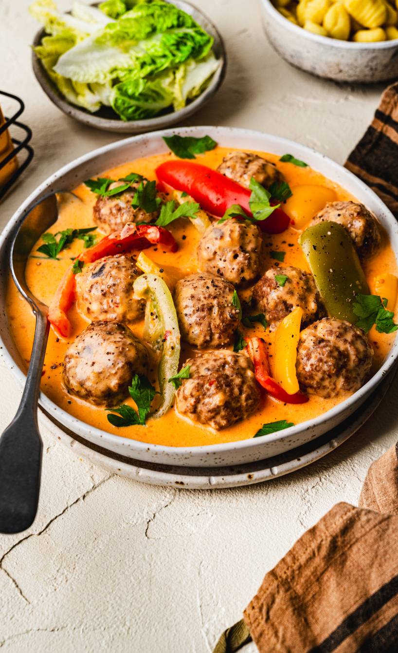 Selbst gemachte Hackbällchen in sahniger Paprikasoße auf einem tiefen Teller, daneben Salat und Gnocchi