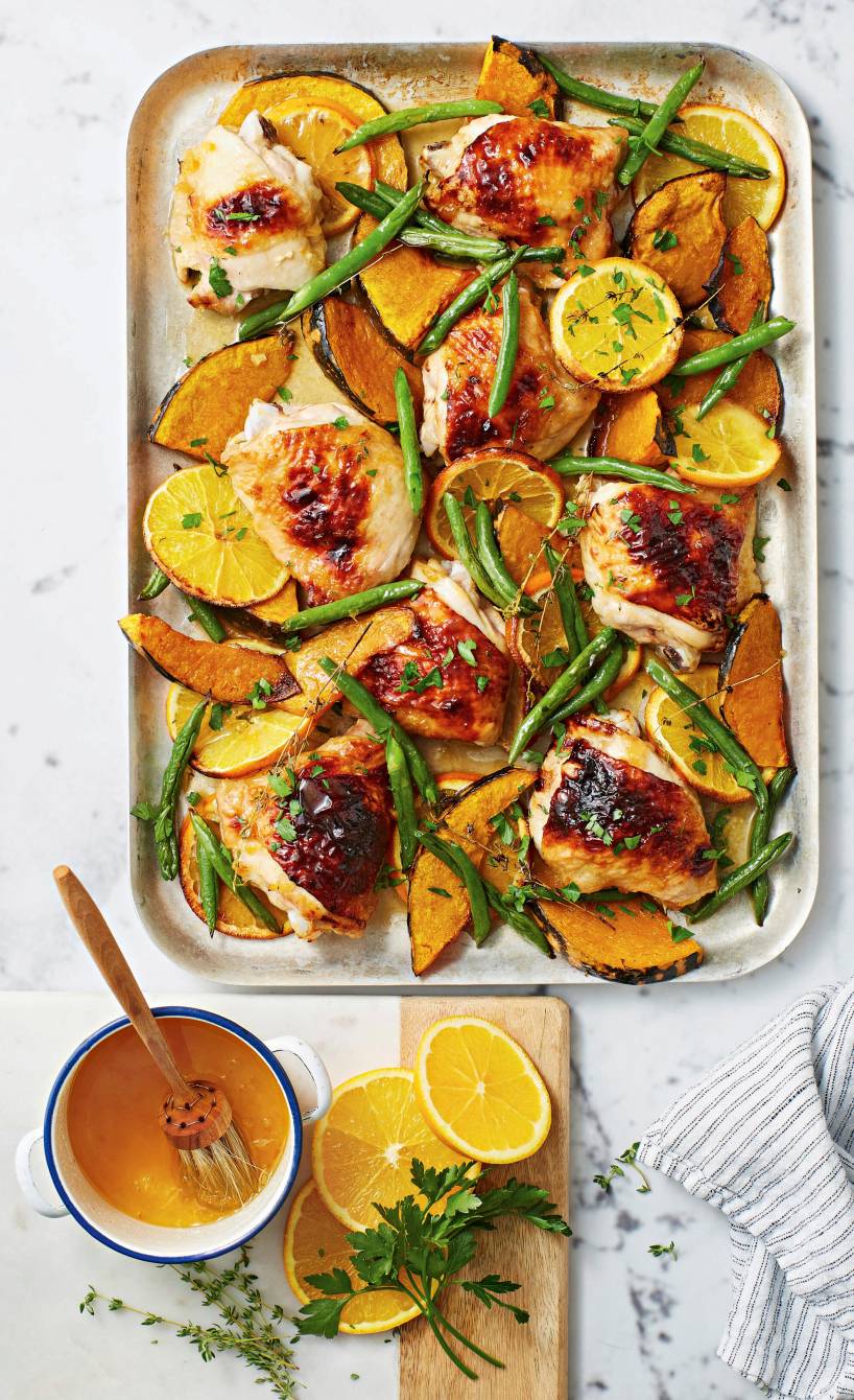 Backblech mit Hähnchenkeulen, Kürbisspalten, grünen Bohnen und Orangenscheiben auf einem Tisch