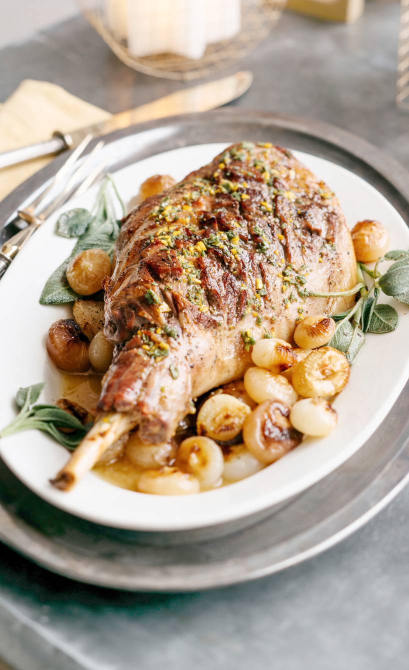 Eine gebratene Lammkeule bestrichen mit mit Senf-Gremolata auf Perlzwiebelgemüseangerichtet auf einem Teller