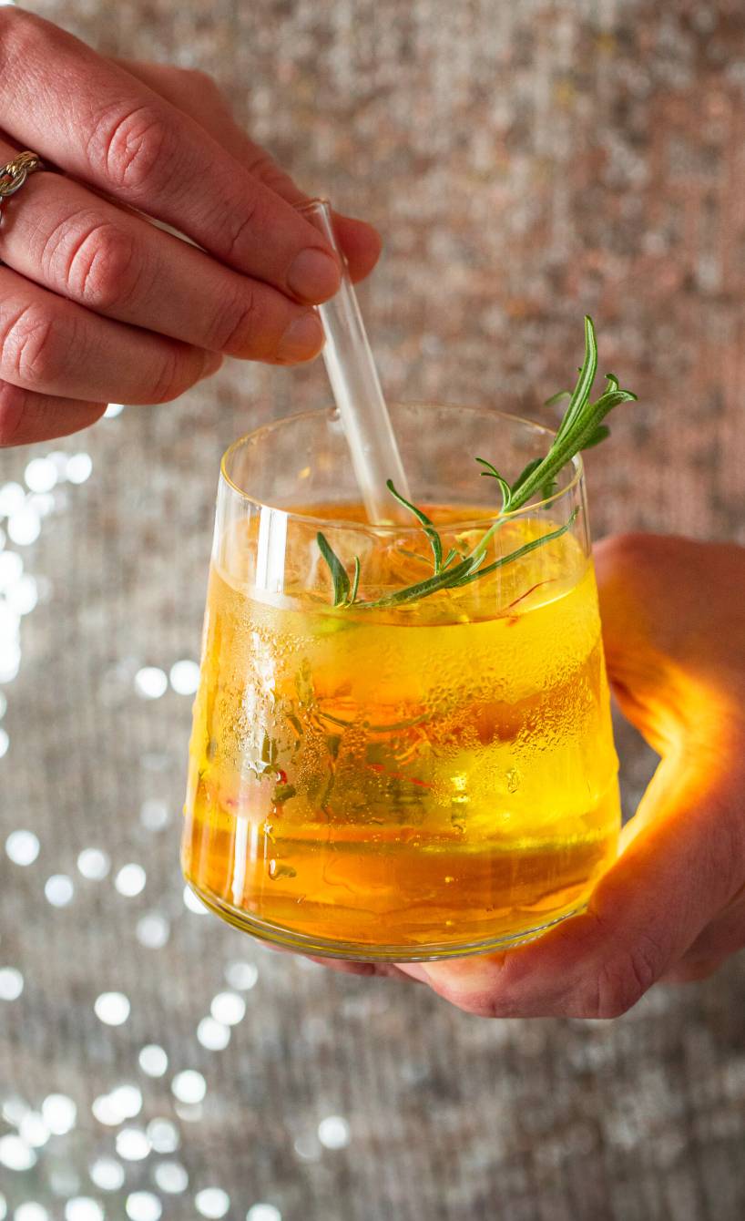 Goldener Gin Tonic mit Safran garniert mit einem Rosmarinzwei in einem Gin Tonic Glas in der Hand einer Frau mit einem Pailettenoberteil