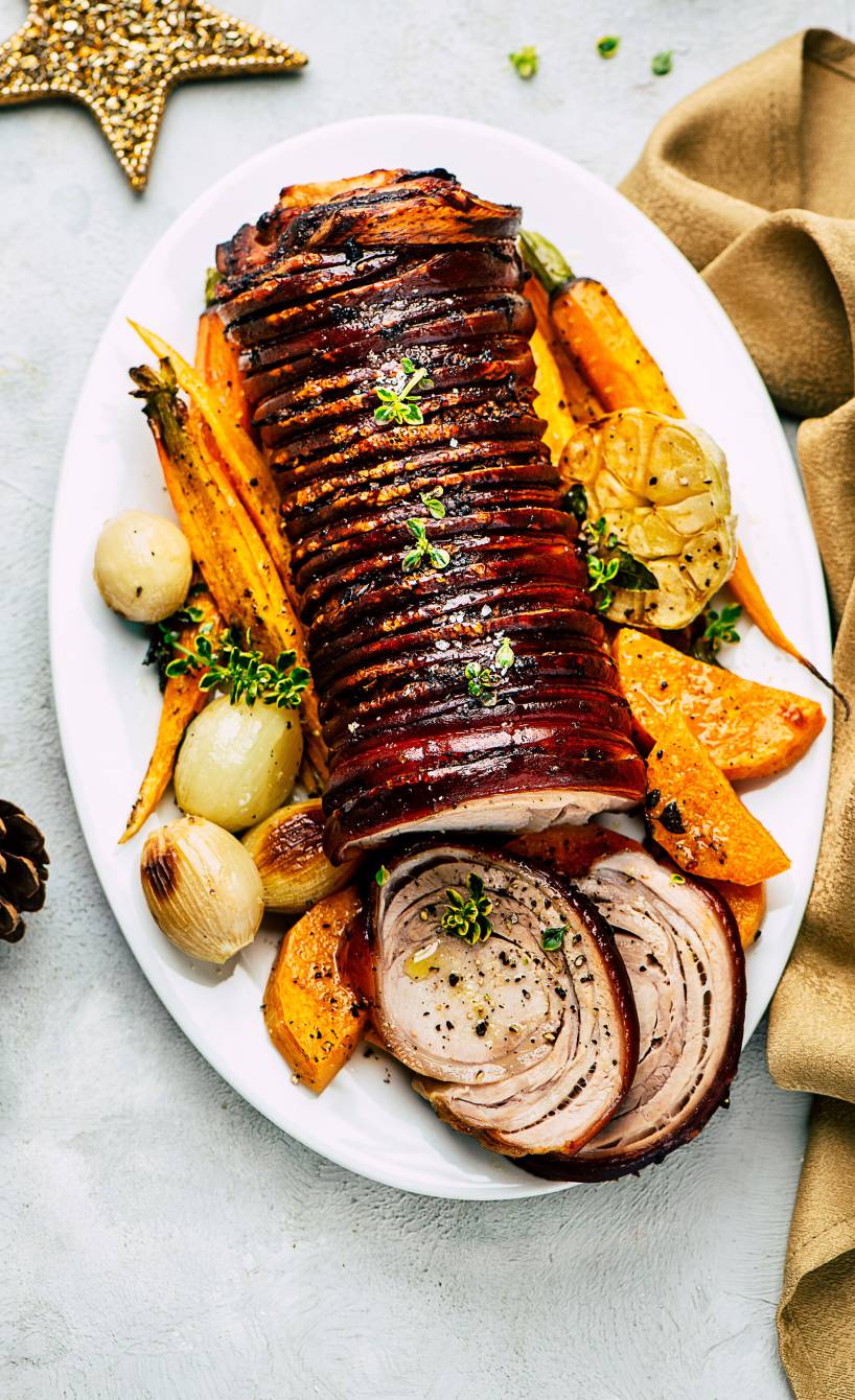 Schweinrollbraten angeschnitten auf einer Servierplatte, drumherum gebackene Süßkartoffeln und Karotten