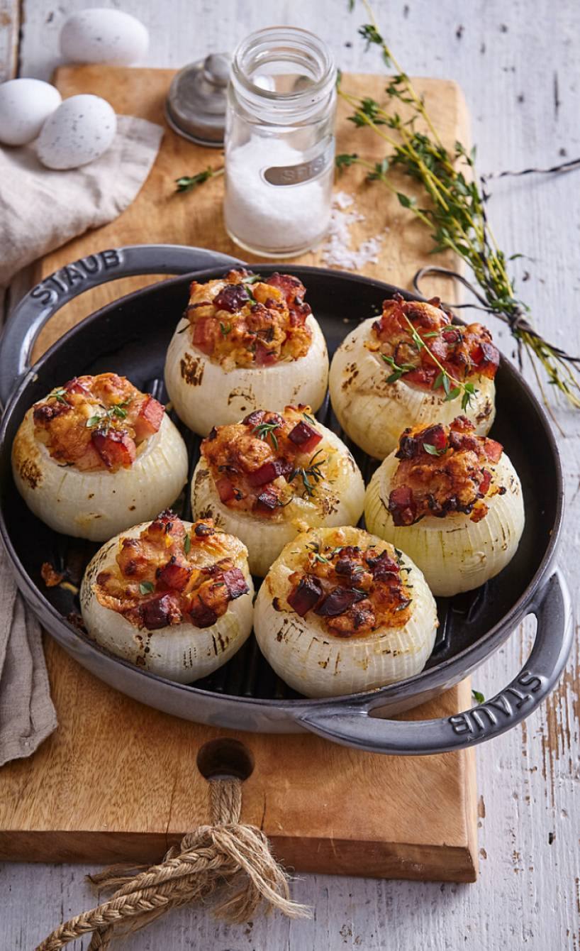 gebackene, gefüllte Zwiebeln mit Pancetta, Brot und Hüttenkäse in einer Ofenform auf einem Holztisch