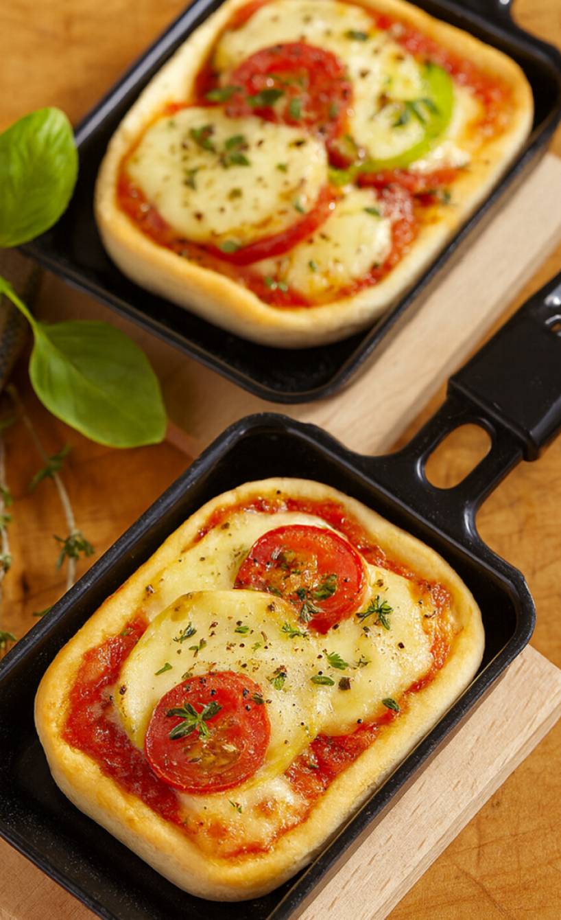 Zwei Raclette-Pfännchen gefüllt wie eine Mini-Pizza mit Teig, Tomate und Mozzarella