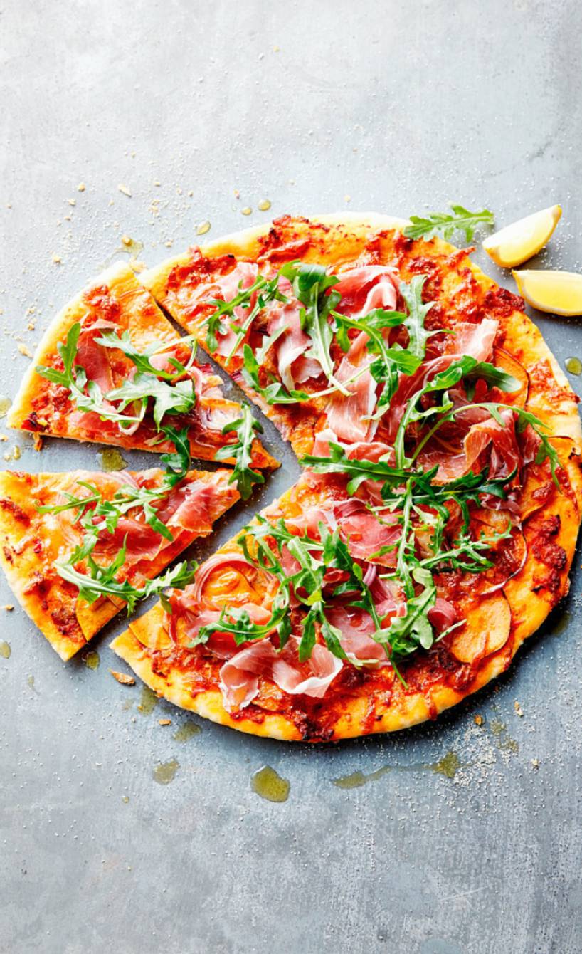 Runde Pizza mit rotem Pesto, Süßkartoffeln, Schinken und Rucola belegt angeschnitten auf einer Arbeitsplatte