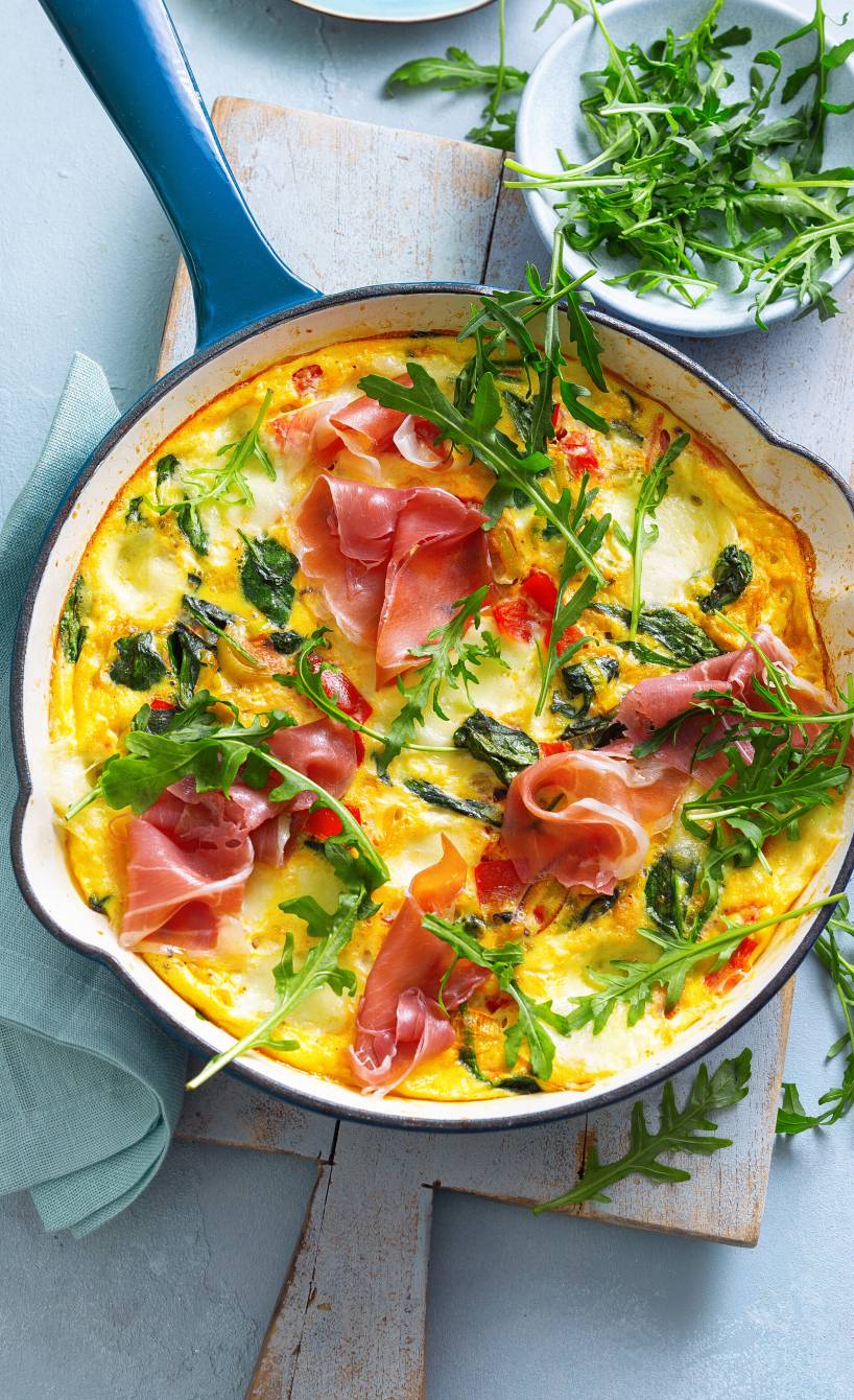 Eine ofenfeste Pfanne mit gebackener Gemüse-Frittata mit Mozzarella und Schinken getoppt von Rucola auf einem Holzbrett