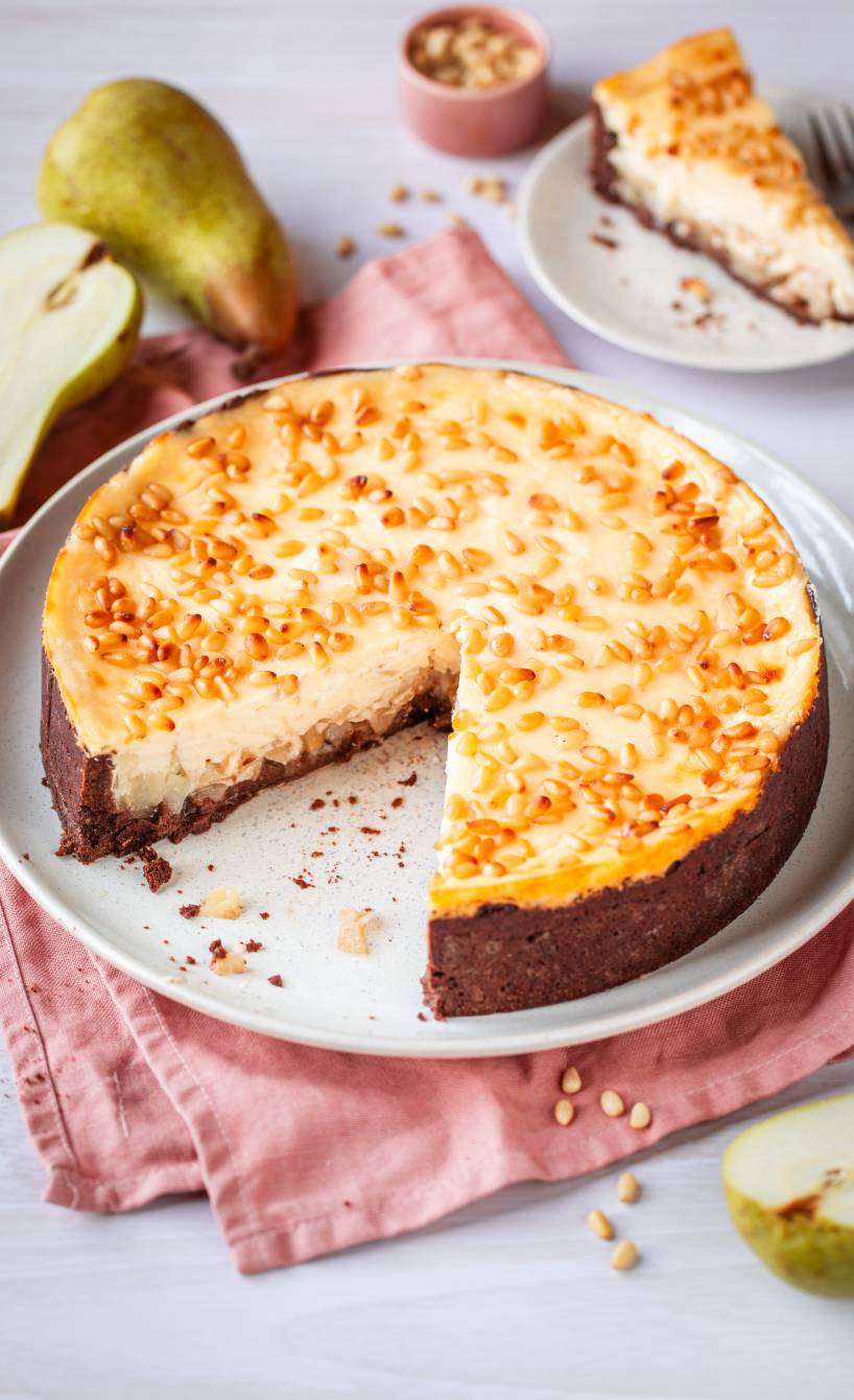 Runder Schmandkuchen mit Birnen, Schokoboden und Pinienkernen angeschnitten auf einer Kuchenplatte