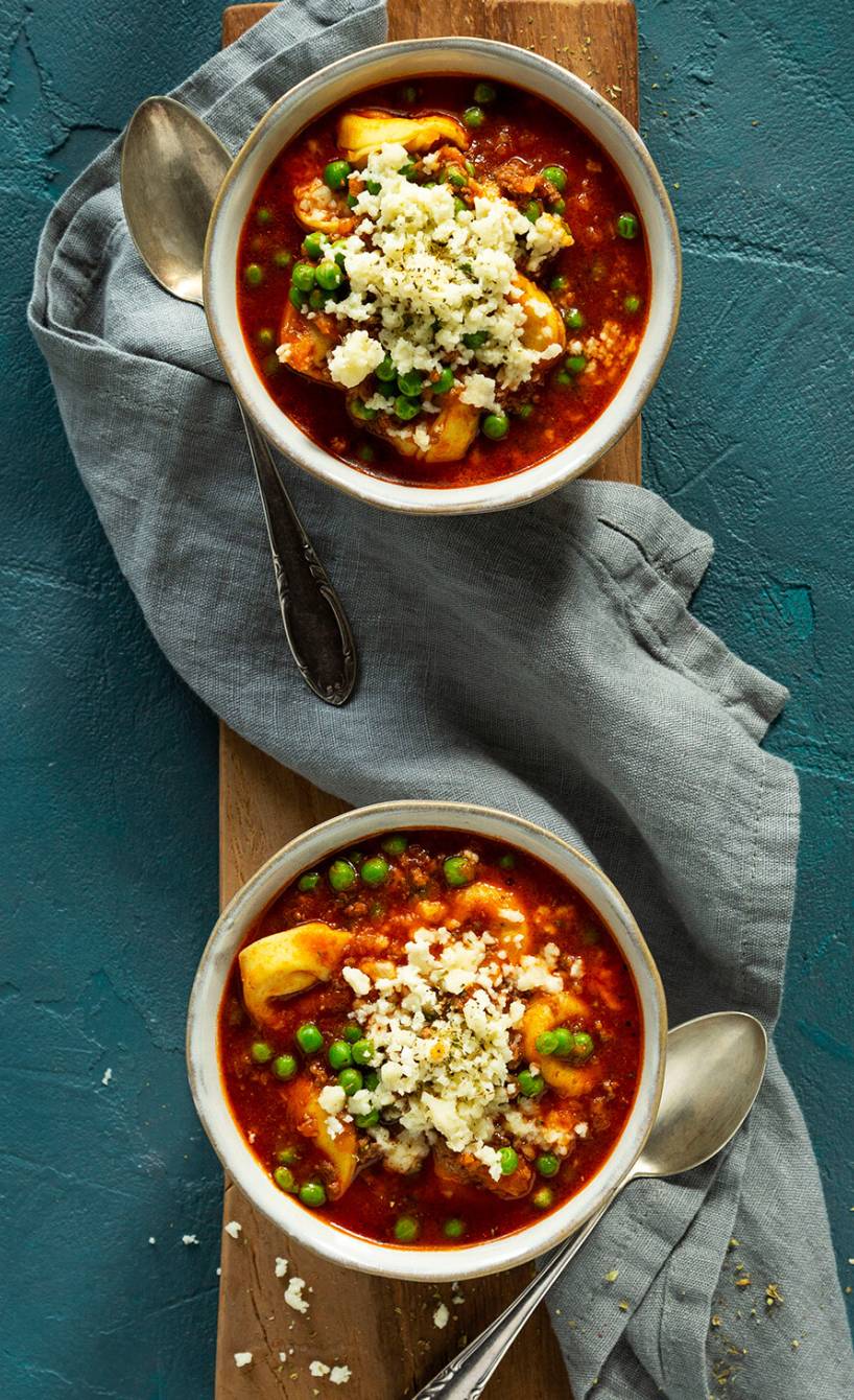 Bolognese-Suppe mit Tortellini und Käsetopping in zwei Suppenschalen auf einem Holzbrett