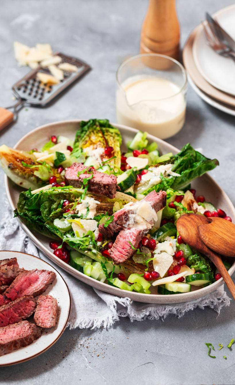 Gebratener Römersalat angerichtet mit Steak-Streifen und Granatapfelkernen auf einer Platte, daneben Parmesan-Joghurt-Dressing