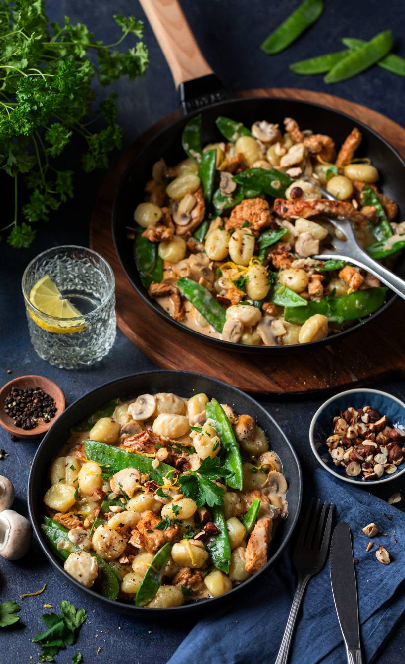 Pfanne mit Gnocchi, Champigons, Zuckerschoten und Putenbruststreifen getoppt von Haselnüssen auf einem Tisch