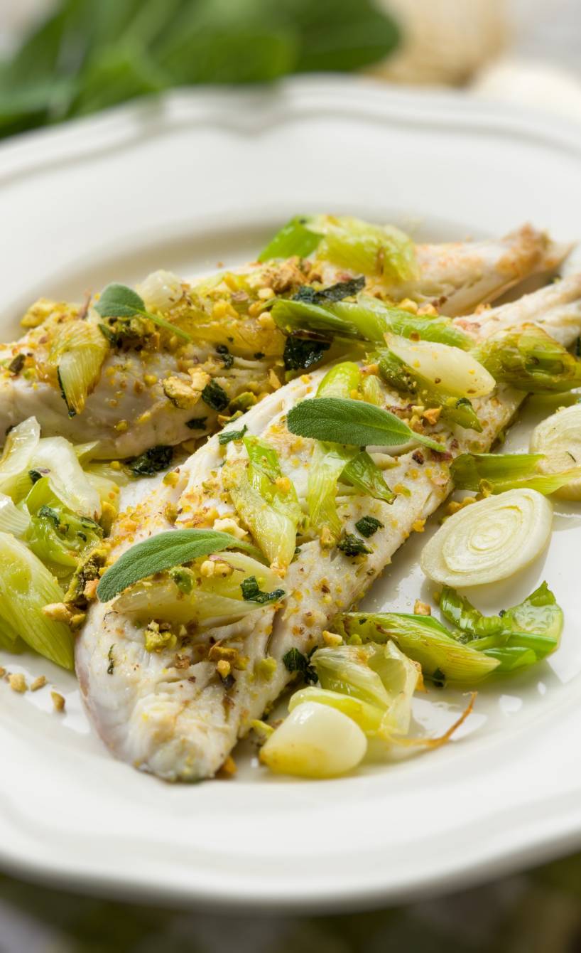 Zwei gebratene Kabeljaufielts auf einem Teller mit Porreeringen und gerösteten Pistazien