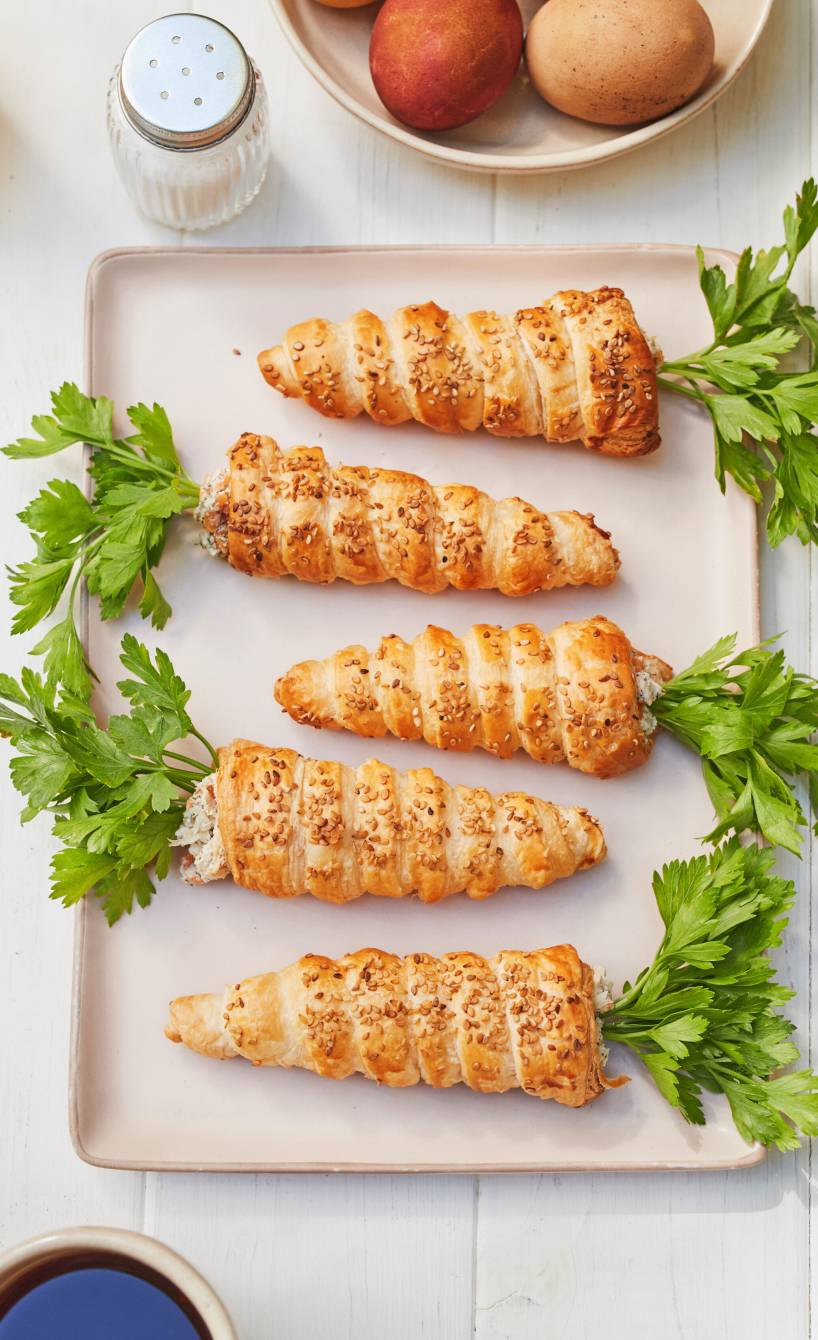 Fünf gebackene, gefüllte Blätterteig-Karotten mit Frischkäse-Lachscreme angerichtet auf einer Servierplatte, daneben Ostereier auf einem Tisch