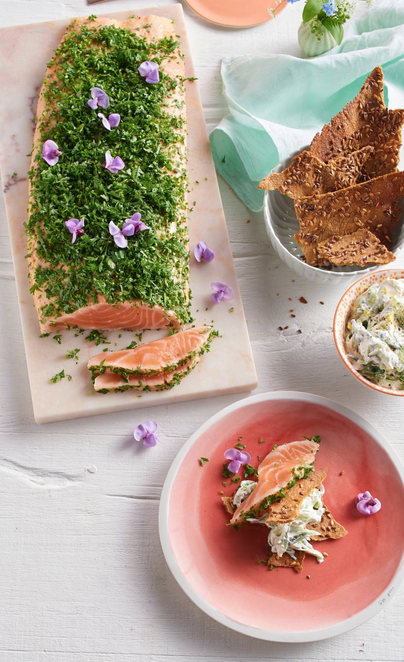 Gebeizte Lachssite im Kräutermantel angeschnitten auf einer Servierplatte, daneben Gurken-Schmand-Dip und Cracker