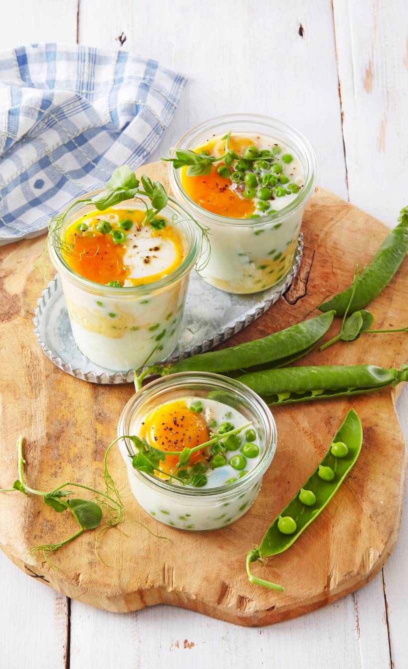 Drei Gläser mit im Ofen gebackenen Eier getoppt von Erbsen-Schalotten-Mix auf einem Holzbrett