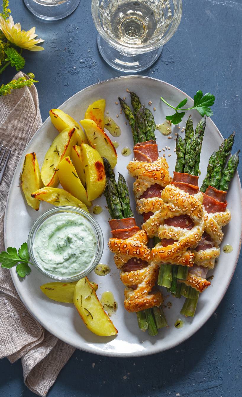 Spargel-Päckchen mit Parmaschinken und Blätterteig angerichtet neben Rosmarin-Kartoffelspalten und Feta-Dip