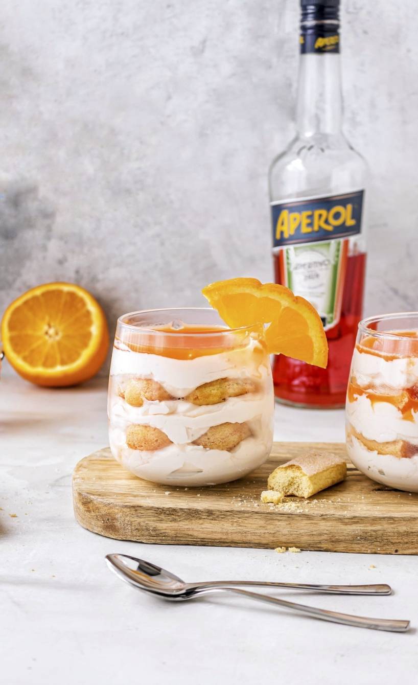 Zwei Gläser mit Aperol Spritzt Tiramisu auf einem Holzbrett, dahinter eine Flasche Aperol