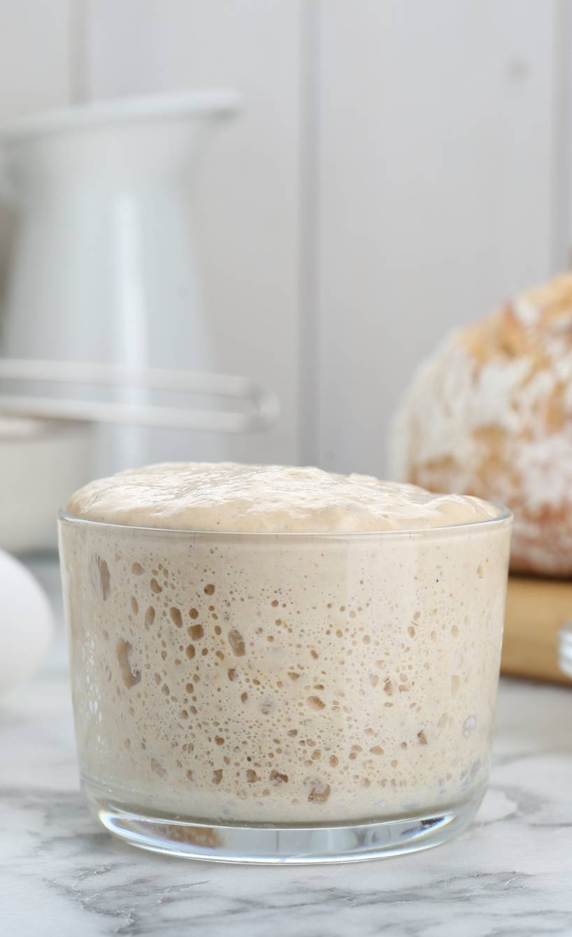 Aktiver Sauerteig in einem Weckglas auf einem Tisch mit einem Sauerteigbrot und Backzutaten