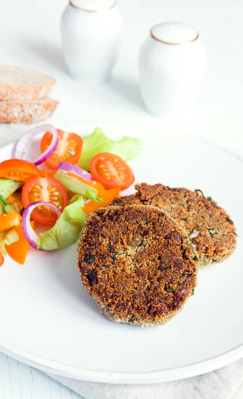 zwei selbstgemachte, gebratene Linsenfrikadellen angerichtet neben gemischtem Salat auf einem Teller