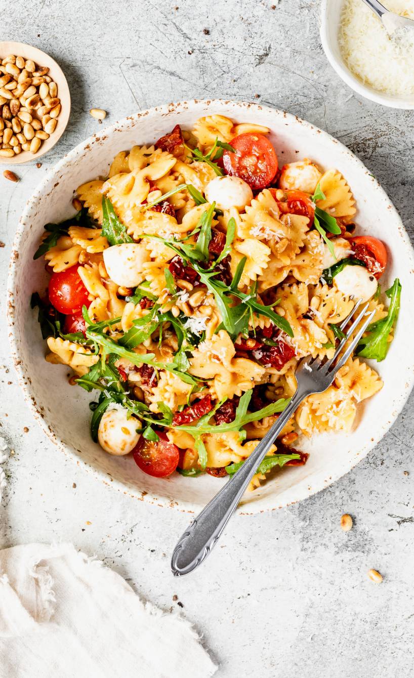 Nudelsalat mit getrockneten Tomaten und Mozzarella angerichtet mit Rucola und Pinienkernen auf einem tiefen Teller