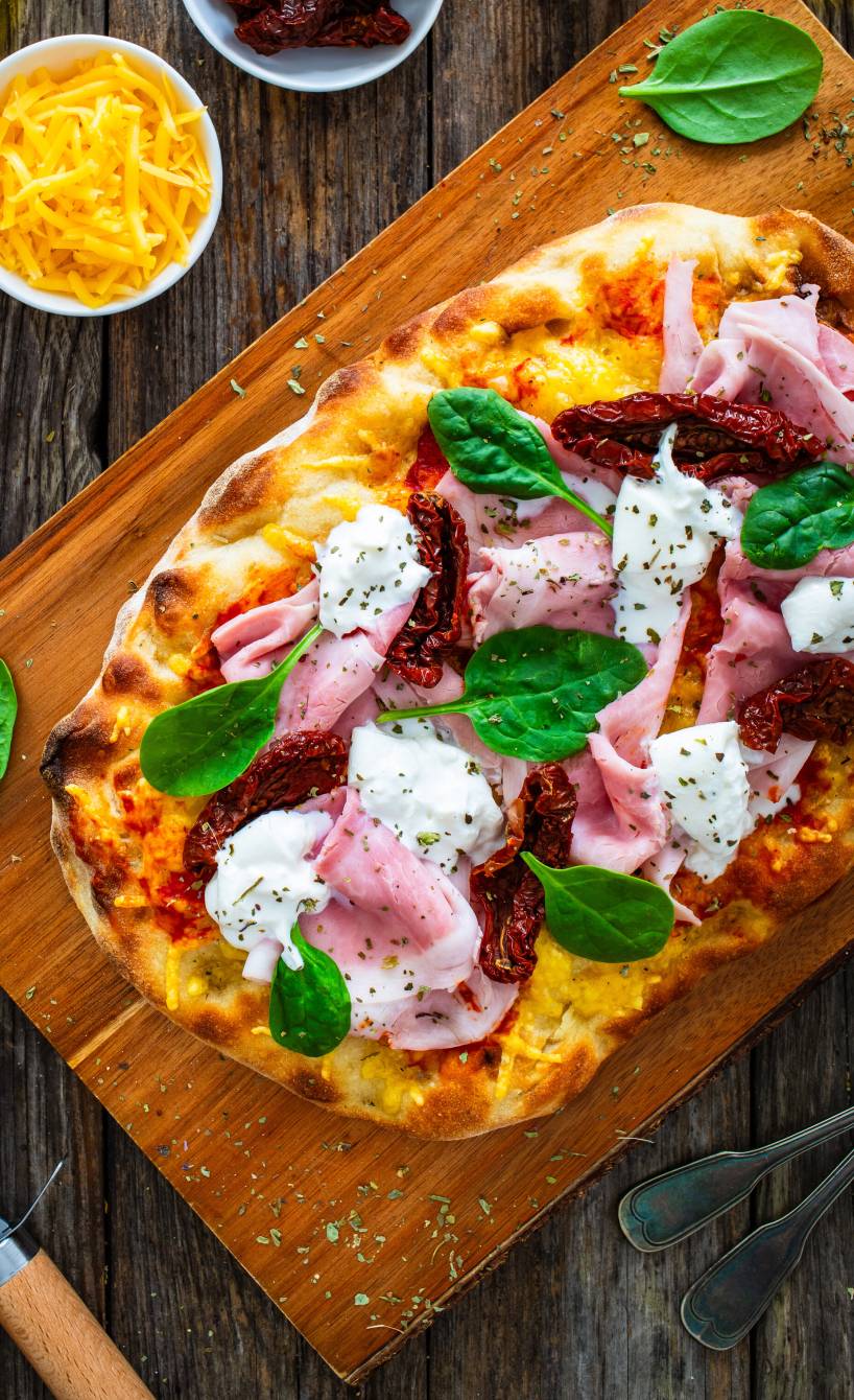 Gebackene, ovale Pinsa belegt mit getrockneten Tomaten, gekochtem Schinken und Burrata auf einem Holzbrett