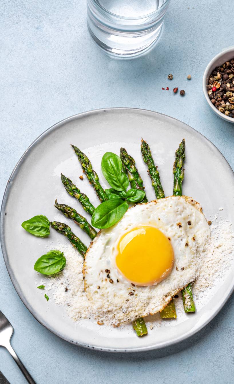 Spargel auf Mailänder Art mit Spiegelei und Parmesan angerichtet auf einem Teller getoppt von Basilikumblättern