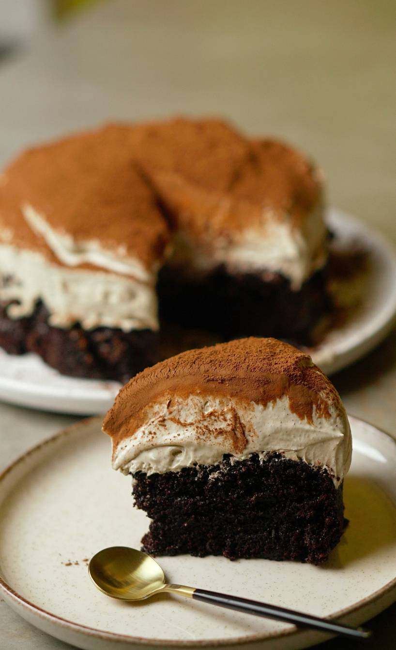 Ein Stück Tiramisu-Kuchen auf einem Kuchenteller, dahinter der Kuchen auf einer Tortenplatte