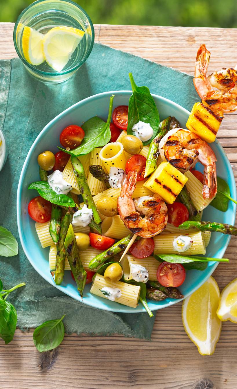 Nudelsalat mit gegrilltem Spargel getoppt von einem Garnelen-Mango-Spieß in einer Salatschüssel auf einem Gartentisch, daneben Knoblauch-Joghurt-Dressing