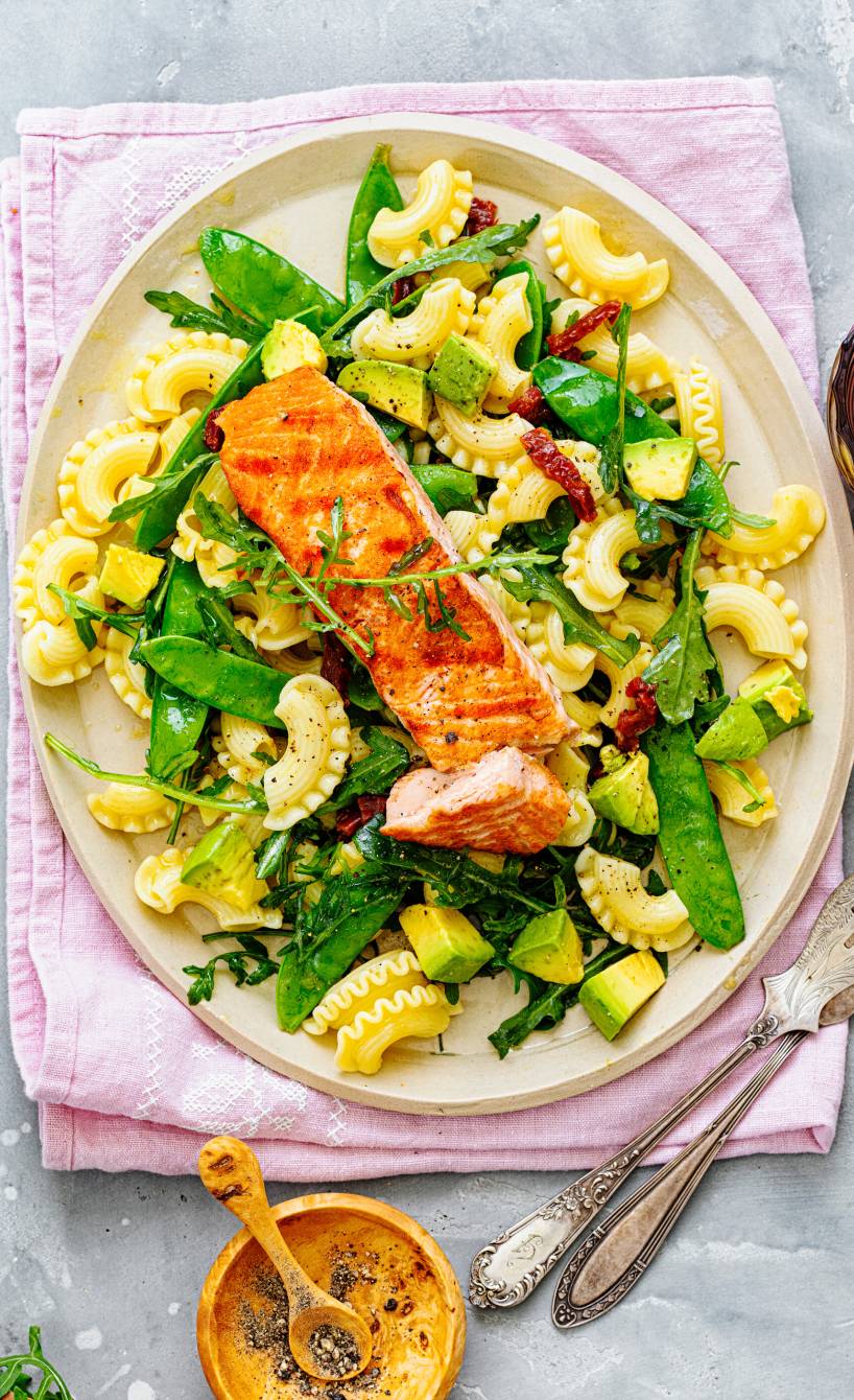 Nudelsalat mit Rucola, Avocado, getrockneten Tomaten, Zuckerschoten angerichtet mit einem Stück gegrilltem Lachsfilet auf einem Pastateller