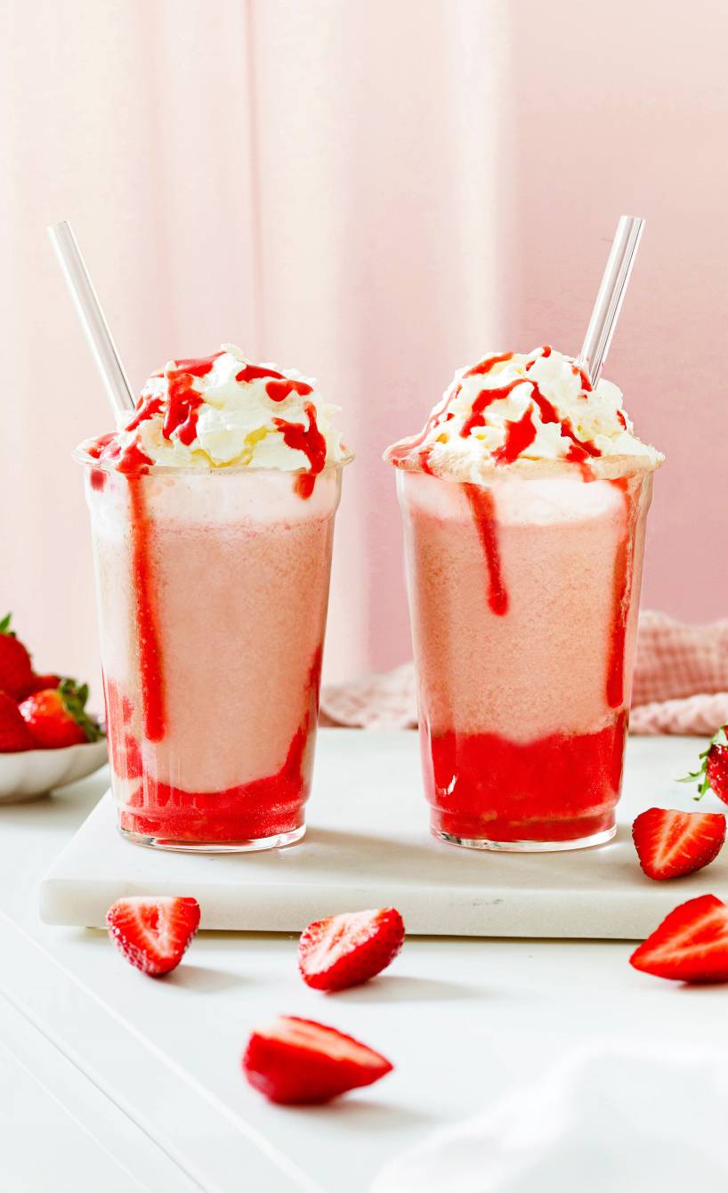 Zwei selbstgemachte Erdbeer-Eiskaffees in hohen Gläsern getoppt von Sahne und frischen Erdbeeren auf einem Tisch
