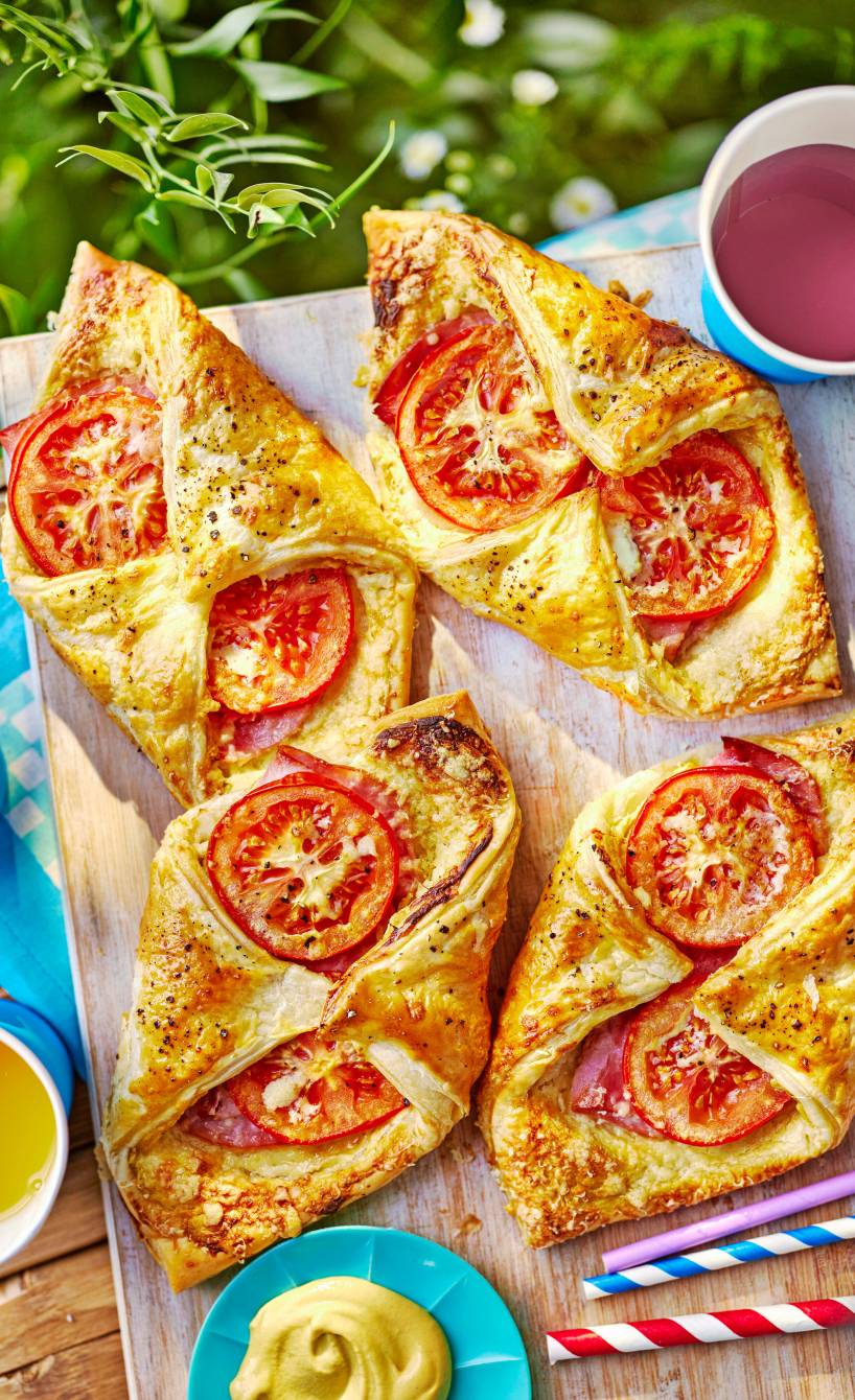 Vier selbstgebackene Schinken-Käse-Blätterteigtaschen mit Tomaten auf einem Holzbrett im Garten