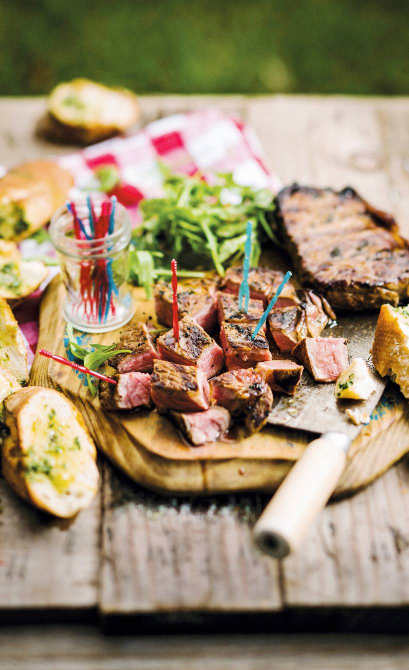 Gegrillte Rindersteaks angerichtet mit selbstgemachtem Knoblauch-Ciabatta und Rucola auf einem Holzbrett im Garten