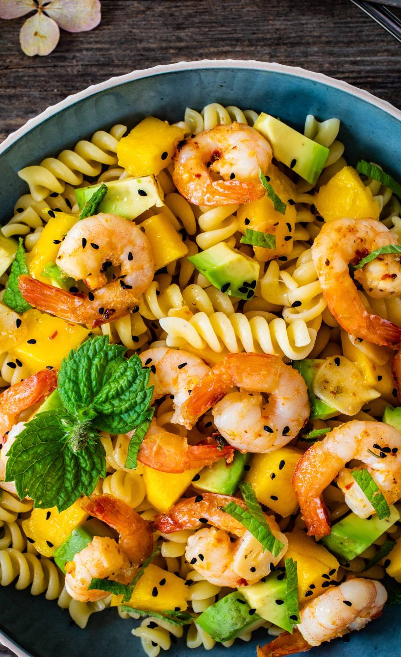 Nudelpfanne aus Fusilli mit Garnelen, Mango, Avocado getoppt von frischer Minze auf einem Tisch