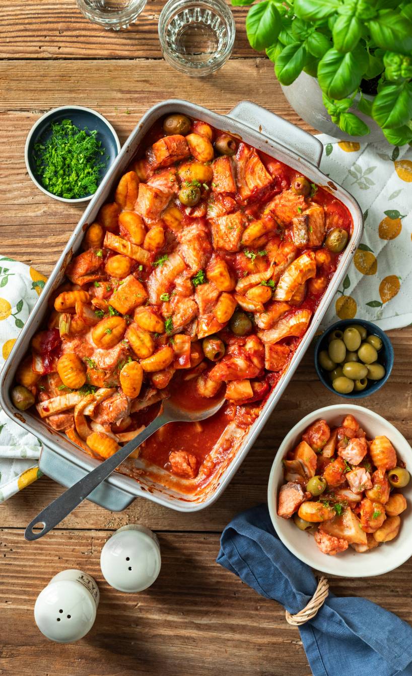 Lachs-Gnocchi-Auflauf mit Fenchel und Tomatensoße auf einem Holztisch, daneben grüne Oliven und Basilikum