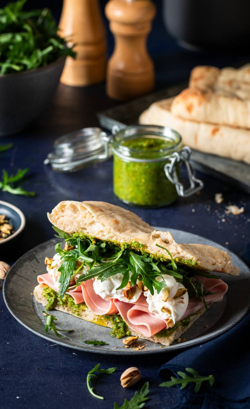 Ein gefülltes Pinsa-Panino mit Mortadella, Burrata und Rucola angerichtet mit selbstgemachtem Pisatzienpesto auf einem Teller