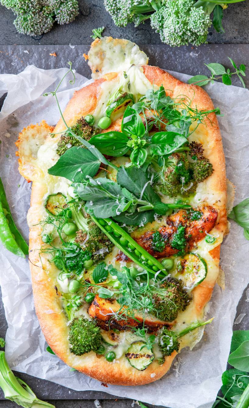 Selbstgebackene, ovale Pizza Bianca belegt mit Hähnchen, grünem Gemüse und Ricotta-Creme getoppt von grünem Pesto auf einer Schieferplatte