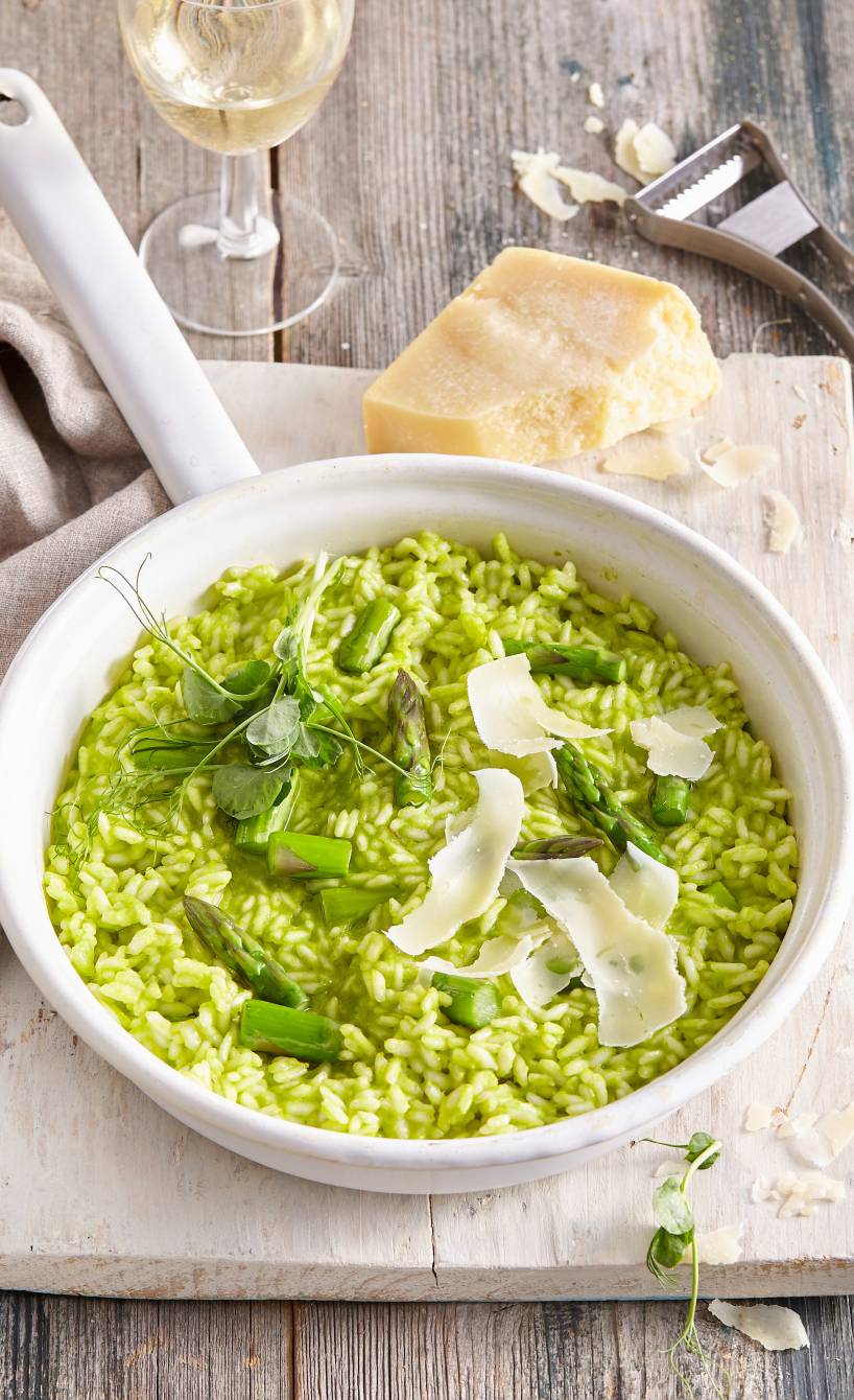 Risotto mit grünem Spargel auf einem tiefen Teller getoppt von Parmesanspänen auf einem Holztisch, daneben ein Glas Weißwein