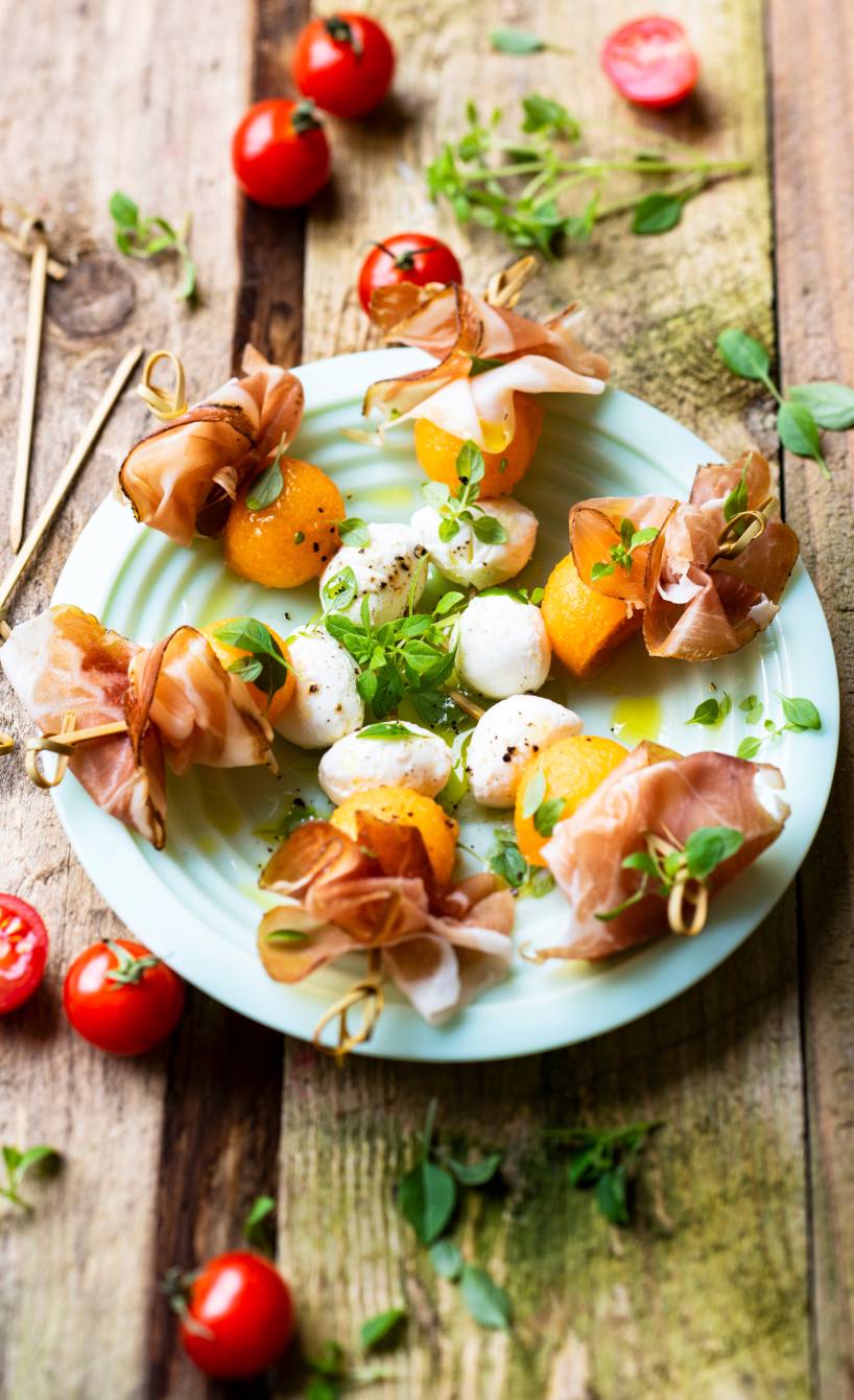 Mehrere Melone-Mozzarella-Spieße mit Schinken angerichtet auf einem Vorspeisenteller auf einem Holztisch