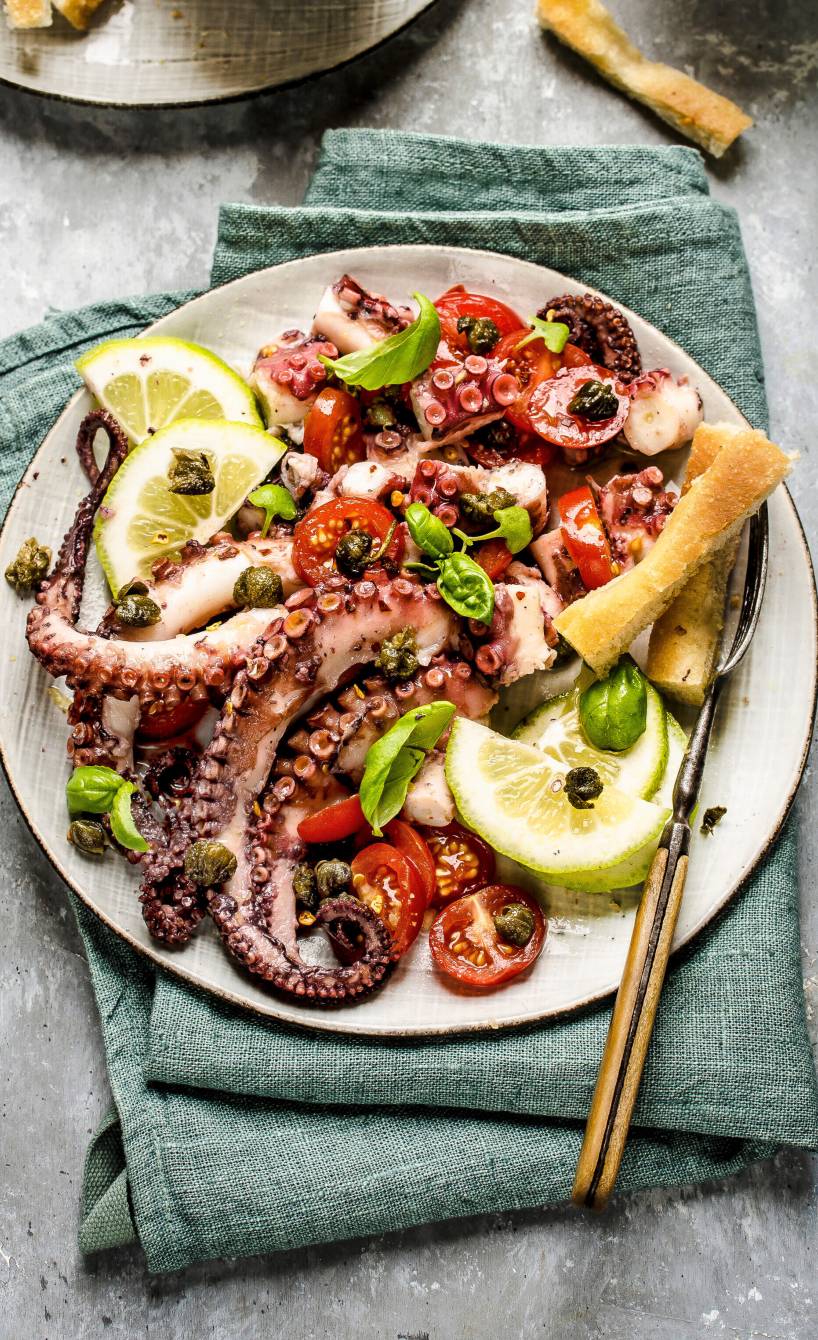 Warmer Oktopussalat angerichtet auf einem Teller mit Zitronen, Basilikum und Focaccia-Streifen