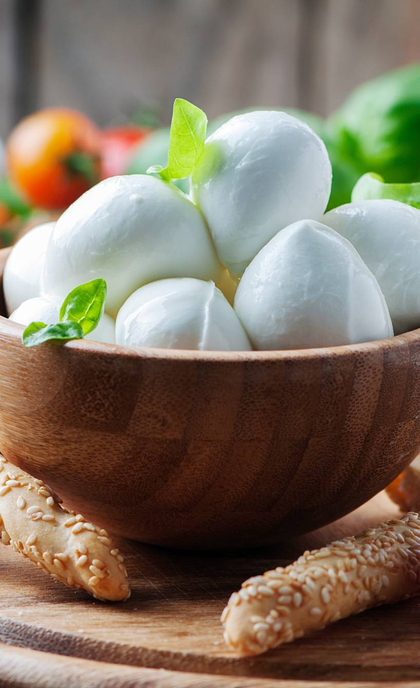 Mozzarellakugeln in einer Holzschale umgeben von Tomaten, Grissini und Basilikum