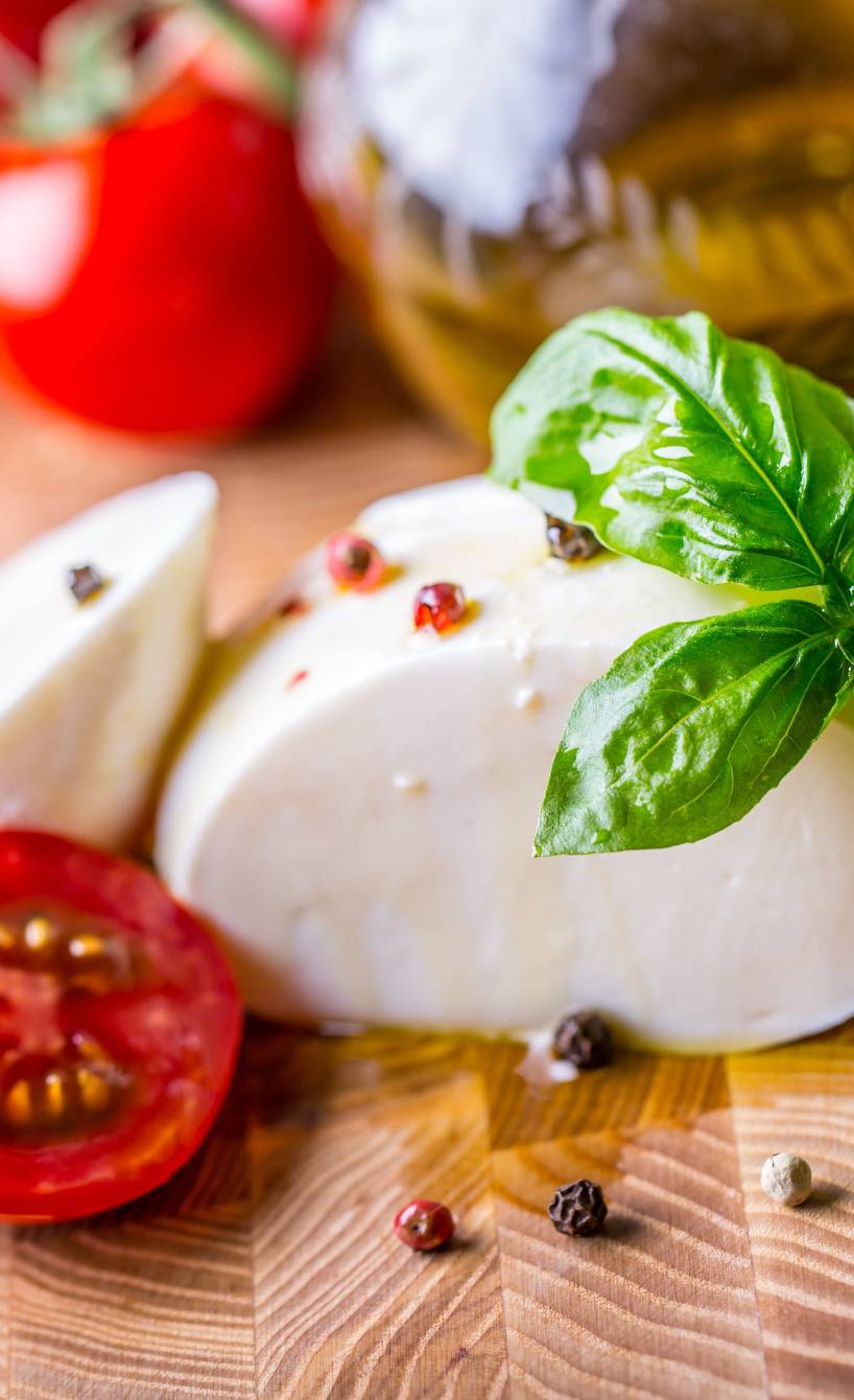 Eine angeschnittene Mozzarellakugel auf einem Holzbrett, daneben Strauchtomaten, Basilikum, Olivenöl und Pfefferkörner