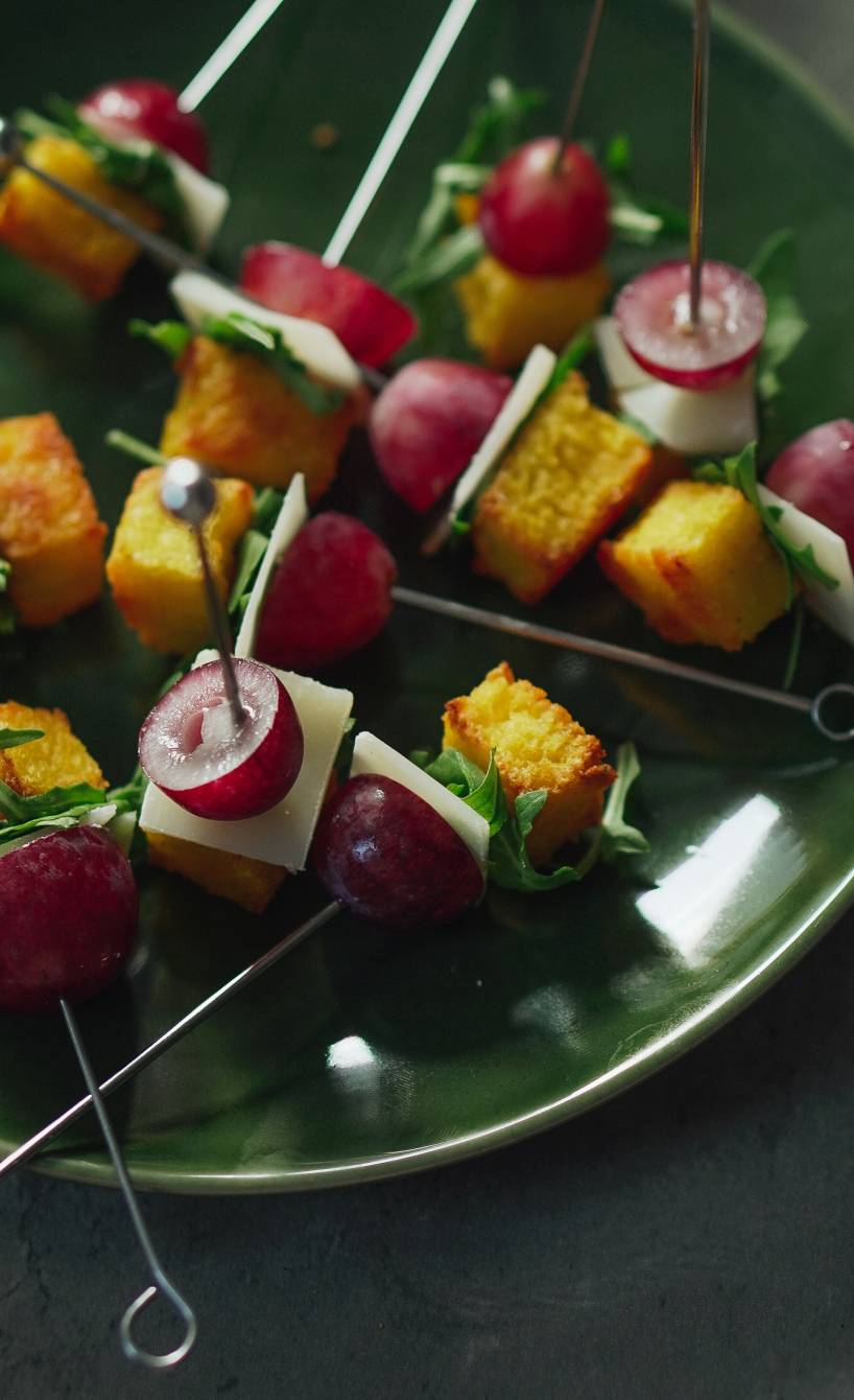 Parmesan-Polenta-Häppchen mit Rucola und Trauben auf Spießen angerichtet auf einem Teller