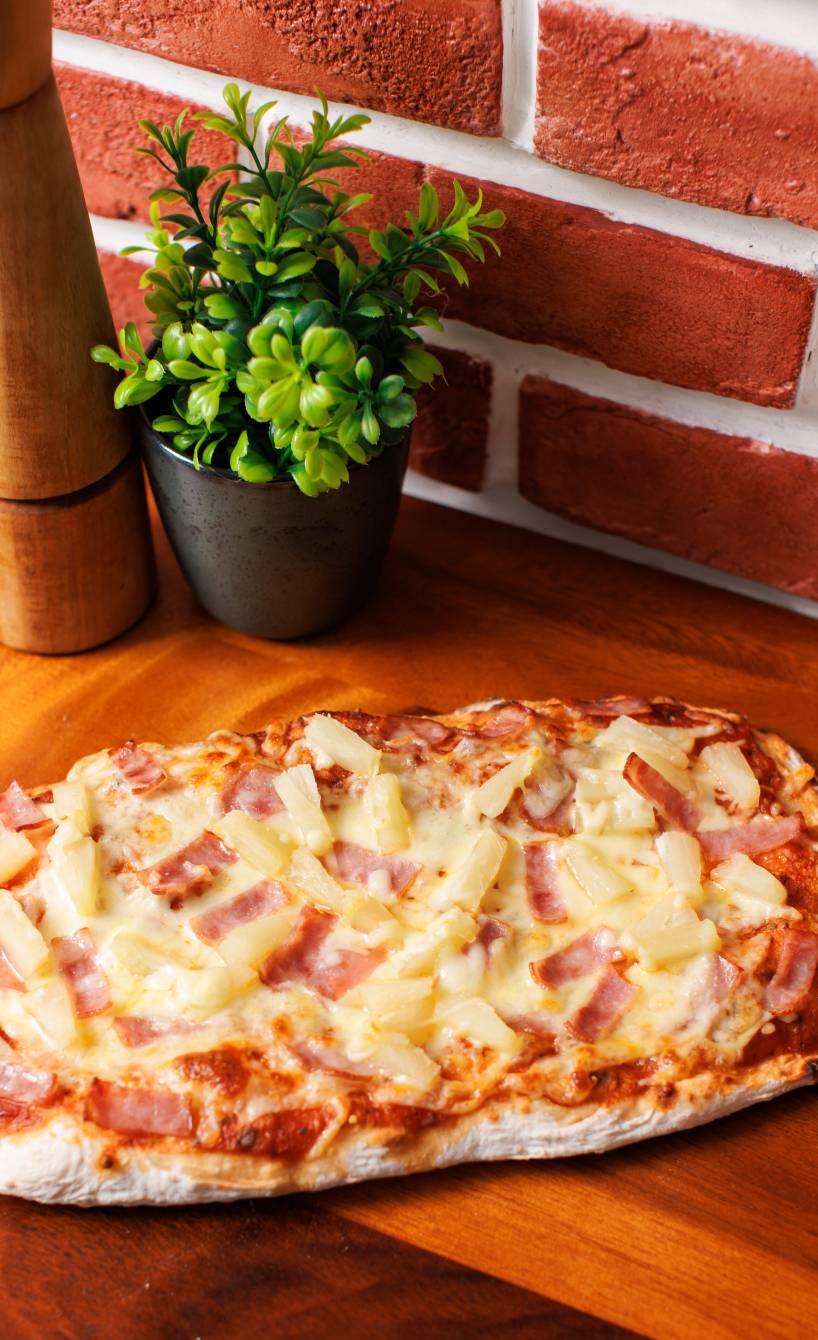 Eine gebackene Pinsa Hawaii mit Mozzarella, Schinken und Ananas auf einem Holzbrett