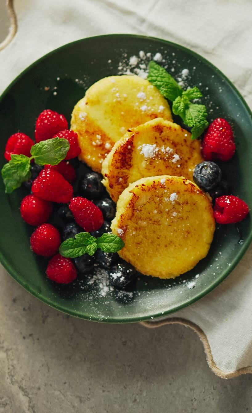 Drei gebratene Polenta-Pfannkuchen getoppt mit frischen Beeren, Puderzucker und Ahornsirup sowie frischer Minze auf einem Teller
