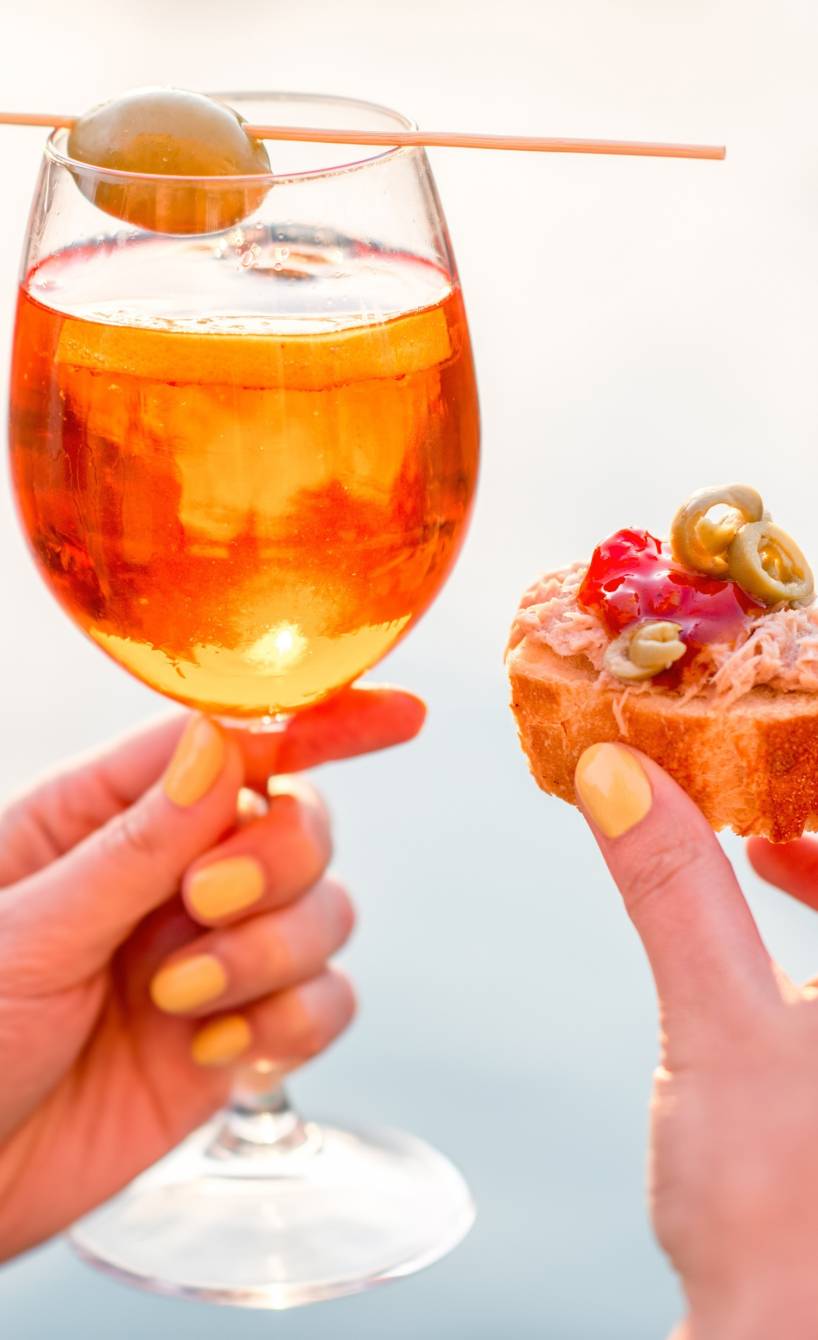 Eine Frau hält in der einen Hand einen Aperol Spritz und in der anderen ein Bruschetta zum Aperitif