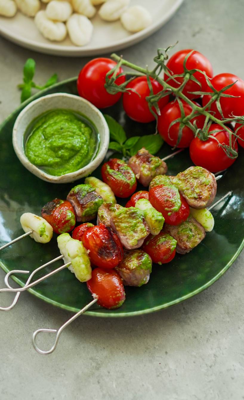 Gegrillte Gnocchi-Salsiccia-Spieße angerichtet auf einem Teller mit Pesto und frischen Tomaten