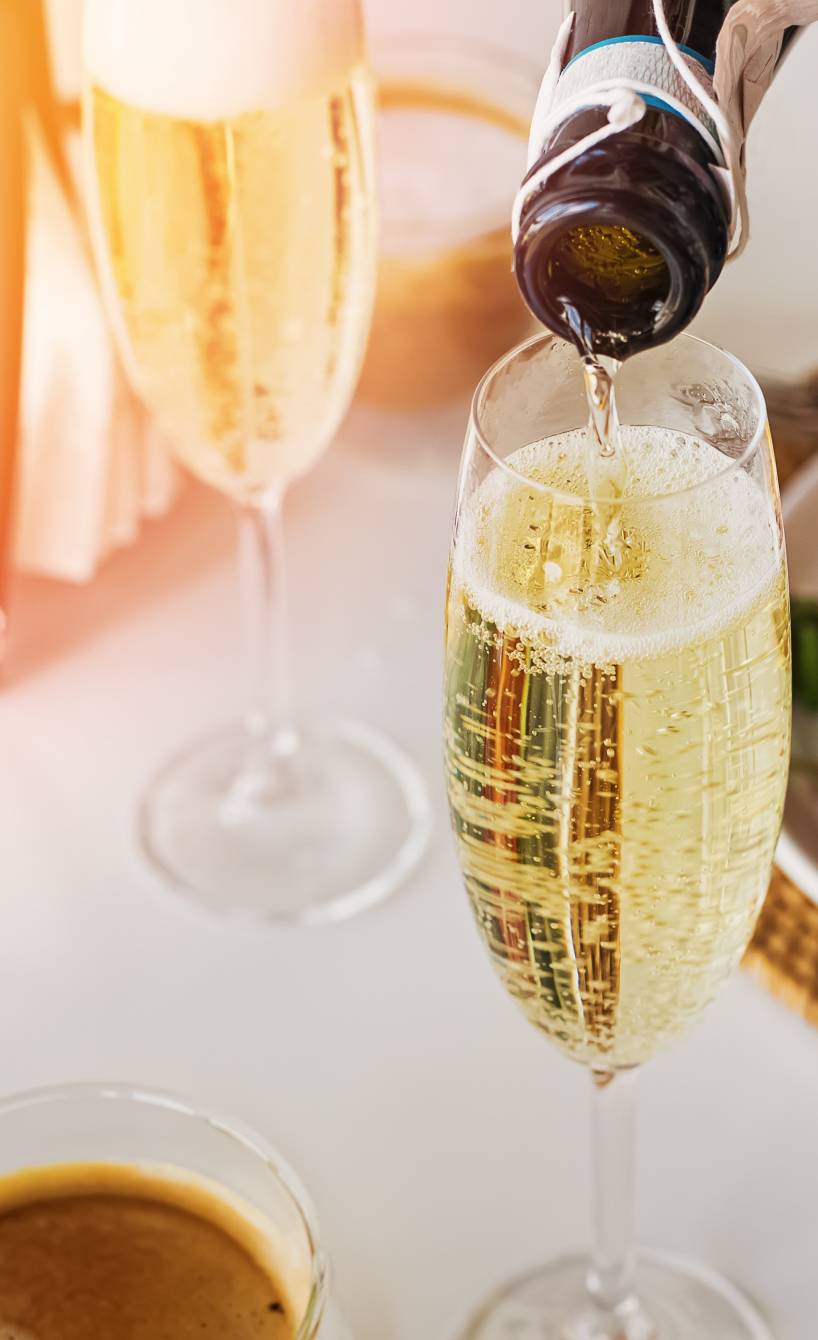 Prosecco wird in ein Glas geschüttet, dahinter ein gedeckter Tisch mit Antipasti Prosecco wird in ein Glas geschüttet, dahinter ein gedeckter Tisch mit Antipasti