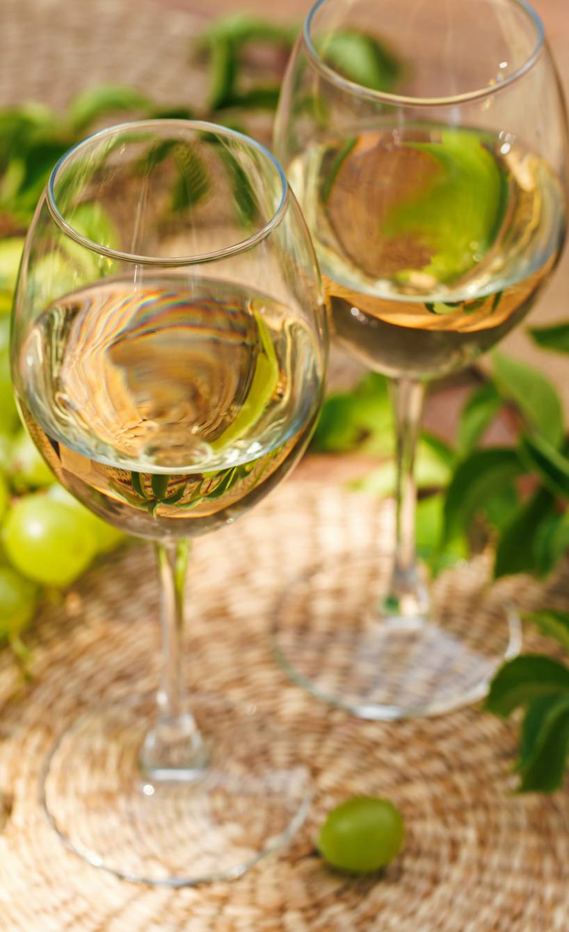 Zwei Gläser mit Weißwein auf einem Gartentisch, daneben grüne Trauben und ein Strohhut Zwei Gläser mit Weißwein auf einem Gartentisch, daneben grüne Trauben und ein Strohhut