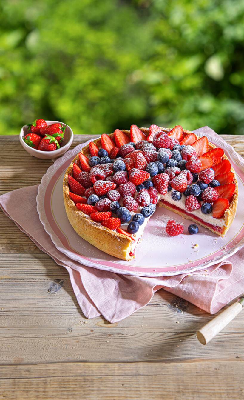 Beerentarte mit Ricotta-Quark-Creme und Johannisbeergelee angeschnitten auf einer Tortenplatte auf einem Holztisch im Garten