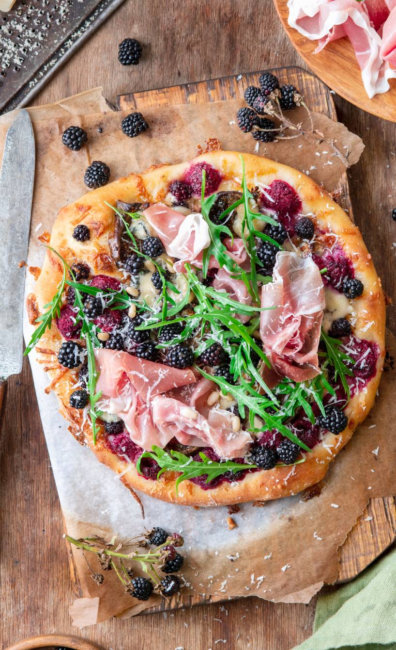 Selbstgebackene Brombeerpizza mit Gorgonzola und Schinken getoppt von Rucola und Pinienkernen auf einem Holzbrett