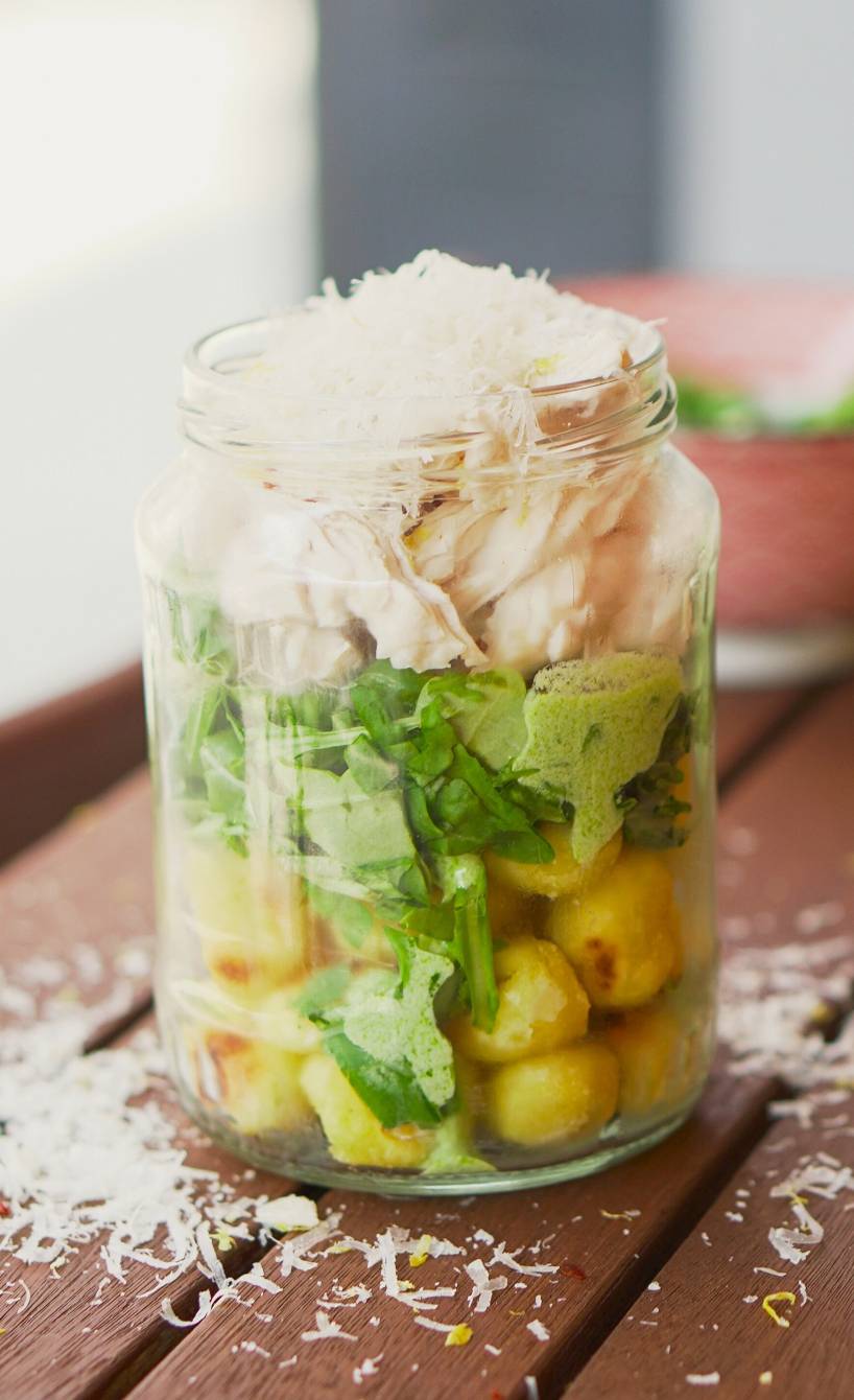 Ein Weckglas als Street Cup mit Schichten aus Gnocchi, Salsa Verde, Rucola und Hähnchen auf einem Holztisch